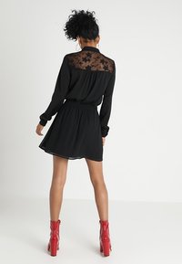 Žena stojící zády k fotoaparátu, oblečená v černých šatech s průsvitnými květinovými krajkovými zády a červenými kotníkovými botami na bílém pozadí.