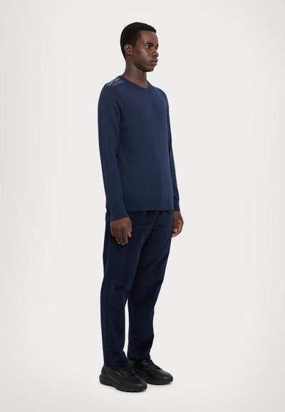 Maglione blu navy con maniche lunghe e scollatura rotonda, con accenti multicolori sulla spalla. Abbinato a pantaloni scuri coordinati e scarpe nere.