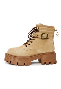 Beige suède enkellaarsjes met een chunky, geribbelde bruine zool, metalen veterogen en een zijgesp. Bruine veters en een trekhaak aan de achterkant.
