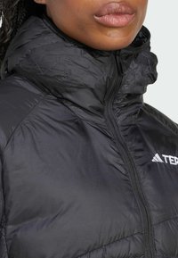 Chaqueta acolchada negra con un alto cuello, cierre de cremallera y un acento de logo. Presenta una textura de tejido suave y brillante.