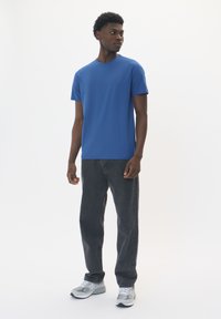 Camiseta azul de manga corta en una tela ligera, combinada con jeans grises y zapatillas grises, de pie contra un fondo blanco.