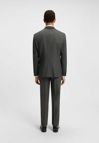 Giacca da abito grigio scuro con una finitura texture, dotata di un'unica fessura sul retro, abbinata a pantaloni dritti coordinati e scarpe nere.