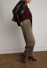 Braune Lederjacke, bordeauxroter Pullover, olivgrüne weit geschnittene Jeans und dunkelviolette High Heels auf einem Holzboden.