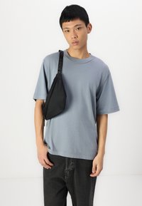 Camiseta de algodón azul claro con mangas cortas, combinada con un bolso cruzado de tela negro. El modelo lleva jeans oscuros; diseño simple y ajuste relajado.