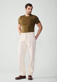 Olajzöld, megfelelően illeszkedő pólót, világos bézs, egyenes szárú nadrágot és barna cipőt viselő férfi, aki háttal kézbe helyezve áll egy szürke háttér előtt.