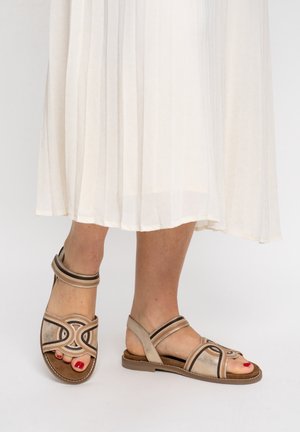 Füße in beigen und braunen flachen Sandalen mit Knöchelriemen, kombiniert mit einem weißen plissierten Midirock vor neutralem Hintergrund.