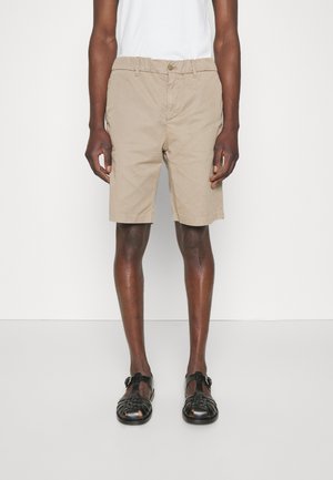Pantaloni corti beige in cotone con frontale piatto, chiusura a bottone e tasche laterali. Indossati con sandali neri intrecciati. Tessuto liscio.