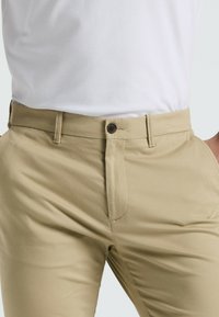 Chinos beiges avec une coupe slim, dotés d'une fermeture éclair, d'un bouton unique, de poches avant et d'une texture de tissu lisse et léger.