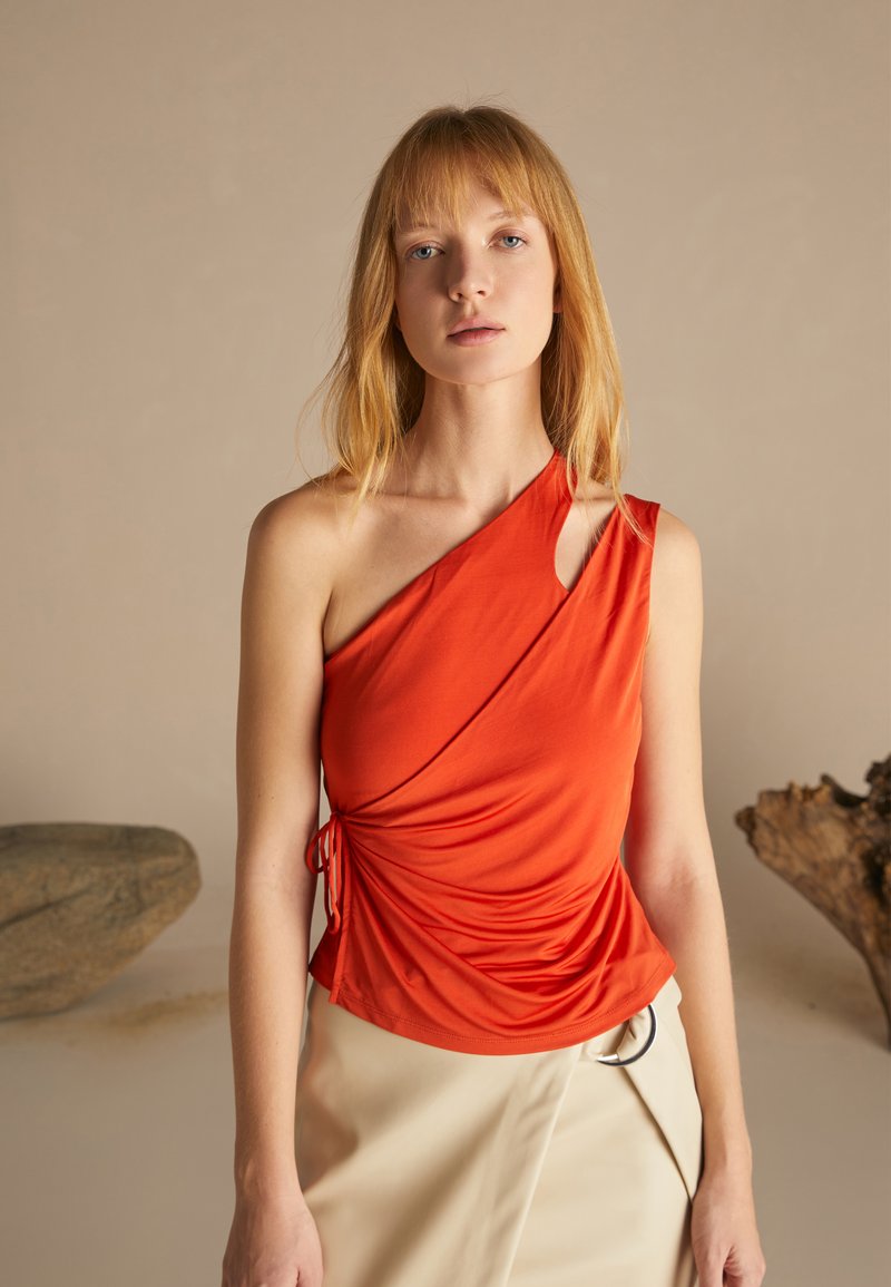 Top de un solo hombro en tela naranja brillante, con textura fruncida y detalle de lazo en el lado, combinado con una falda de color claro.