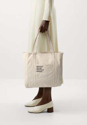 Person i cremefarvet kjole og hvide hæle, der holder en beige tote-taske med teksten "REDUCE REUSE RECYCLE REPEAT" på lommen mod hvid baggrund.