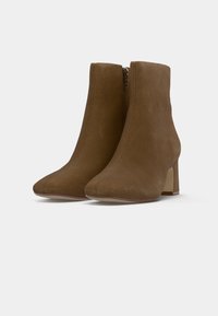 Bottines en daim marron avec bouts pointus, talons blocs et fermetures éclair latérales, présentées sur un fond gris clair uni.