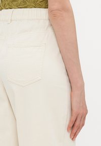 Beige high-waisted trousers with elastic waistband, featuring a single back pocket and smooth cotton texture.