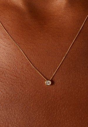 Collier délicat en or avec un pendentif rond serti d'une pierre précieuse claire, reposant sur un fond de teint lisse et foncé.