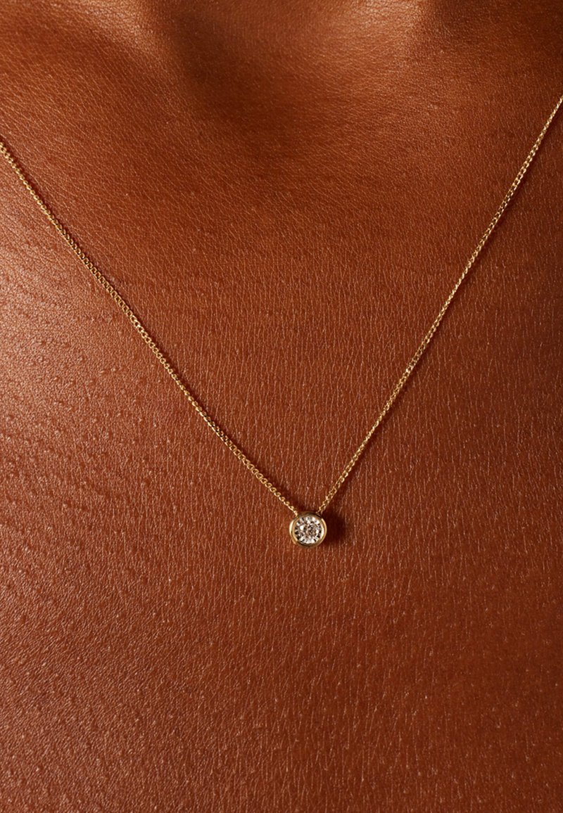 Delicate gouden ketting met een ronde hanger bezet met een heldere edelsteen, rustend tegen een gladde, donkere huidskleur achtergrond.
