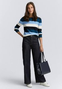 Stripet sweater i blå, sort og hvid; vide, mørke jeans; holder en mørk tote-taske; hvide sportssko. Glatte teksturer og afslappet pasform.