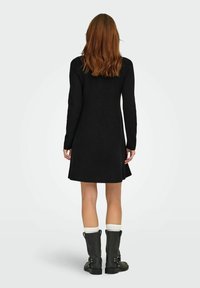 Abito maglione nero dal design svasato con maniche lunghe. Indossato con stivali neri al ginocchio e calze di lana bianche, che mettono in risalto una texture liscia.