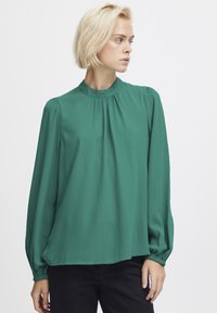 Blusa verde a maniche lunghe con scollo alto, dettagli arricciati sulle spalle, tessuto fluido e polsini elasticizzati. Texture morbida e liscia.