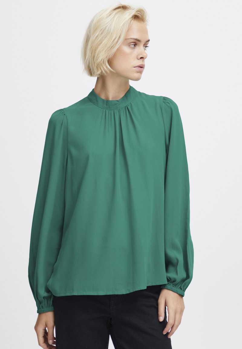Blusa verde a maniche lunghe con scollo alto, dettagli arricciati sulle spalle, tessuto fluido e polsini elasticizzati. Texture morbida e liscia.