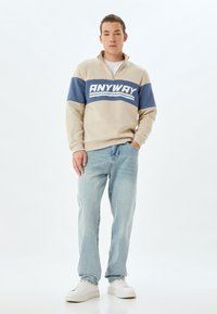 Pull-over beige et marine avec une fermeture éclair quart, arborant un logo blanc "ANYWAY" sur la poitrine, associé à un jean bleu clair et des baskets blanches.