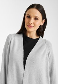Lichtgrijze cardigan van zacht breimateriaal, met een open voorkant en ribbelstructuur, met ingezakte schouders en een relaxte pasvorm.