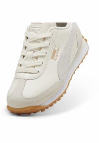 Beige sneaker met glad leer en suède accenten, voorzien van een witte tussenzool, bruine rubberen buitenzool en vetersluiting. Gouden logo detail.