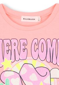 Roze katoenen T-shirt met grote, glinsterende "HIER KOMT" tekst en grafiek van een gestileerd personage met ster- en hartaccenten.