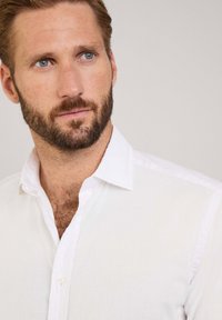 Witte linnen blouse met een knoopkraag en een subtiele textuur, voorzien van een smalle placket en een losse pasvorm. Close-up van het bovenlichaam.