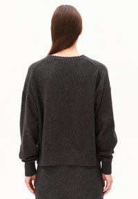 Frau mit langen braunen Haaren, die einen dunkelgrauen Strickpullover und einen passenden Rock trägt, steht mit dem Rücken zu einem weißen Hintergrund.