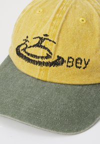 Yellow and green cap with an embroidered design featuring figures and the word "Obey." Made from textured fabric, structured brim.
