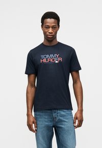 Μπλε ναυτικό βαμβακερό t-shirt με έντονο γράφημα "TOMMY HILFIGER" σε κόκκινο και μπλε. Στρογγυλή λαιμόκοψη, κοντά μανίκια και άνετη εφαρμογή.