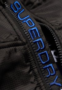 Sort ydre stof med en tekstureret vævning, der har en fremtrædende blå broderet "SUPERDRY"-logo langs lynlåsen.