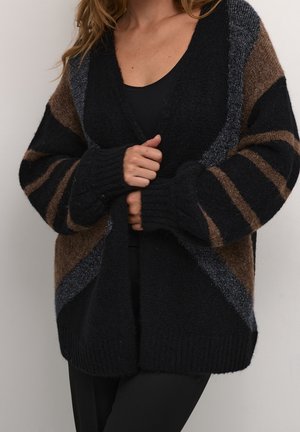 Femme portant un cardigan en grosse maille oversize avec des rayures géométriques noires, marron et grises sur un haut noir, ajustant l'ouverture frontale avec les mains.