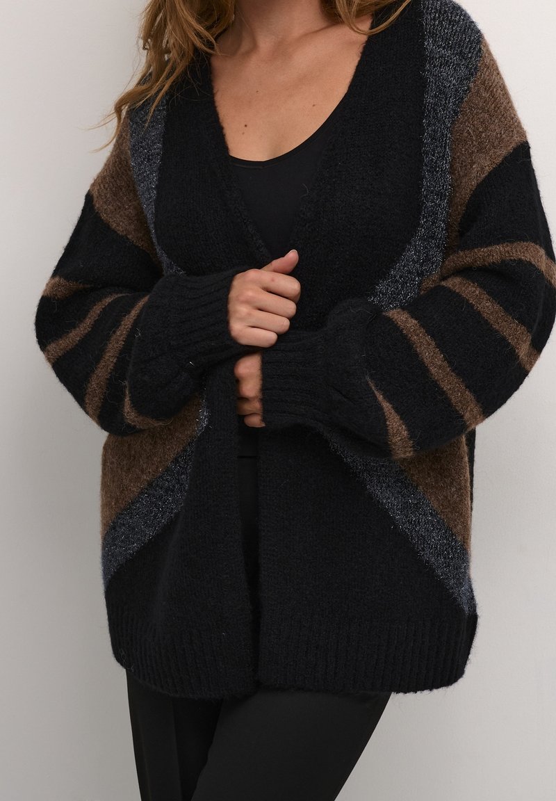 Femme portant un cardigan en grosse maille oversize avec des rayures géométriques noires, marron et grises sur un haut noir, ajustant l'ouverture frontale avec les mains.