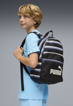 Mochila negra con rayas, cremallera frontal y logo de Puma. Tirantes ajustables y diseño superior curvo. Camiseta azul claro usada debajo.