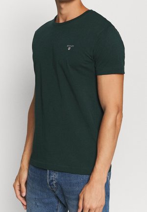 Homme portant un t-shirt vert foncé à manches courtes avec un petit logo GANT sur la poitrine, associé à un jean bleu, debout devant un fond blanc.