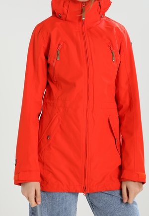 Femme portant une veste à capuche rouge vif avec fermeture éclair avant et poches poitrine zippées, associée à un jean bleu clair.
