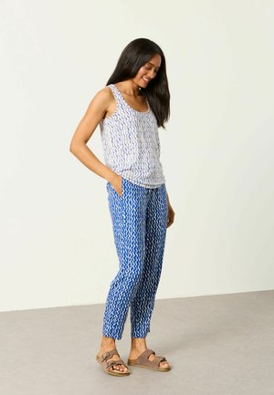 Frau lächelt, steht mit verschränkten Händen, trägt blaue und weiße gemusterte Hose und passendes ärmelloses Oberteil, braune Sandalen auf grauem Boden.