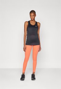 Svart treningstopp uten ermer med rund hals og Nike-logo, sammen med livlige oransje leggings. Modellen har på seg svarte sportsko.