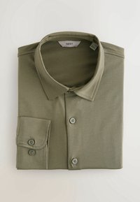 Chemise kaki à manches longues, pliée, avec col et poignets à double bouton sur fond clair.