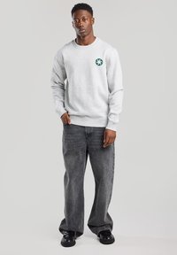 Gray sweatshirt with a circular green logo on the chest, paired with loose-fitting dark gray jeans and black shoes.