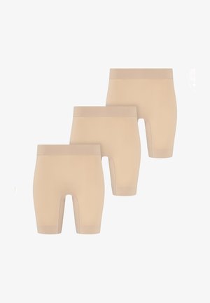 Uśmiechnięta kobieta z upiętymi włosami nosi bezszwowy beżowy biustonosz i wysokie szorty modelujące, stoi boso na jasnym tle.