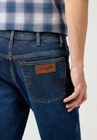 Temno modri denim kavbojke imajo rjav usnjen znak z logotipom "Wrangler", diagonalno šivanje na žepih in ožji kroj.