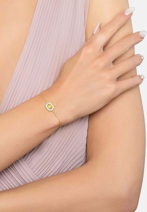 Bracelet en or délicat avec une pierre précieuse jaune encadrée dans un cadre rectangulaire, porté au poignet d'une personne aux ongles manucurés et à la robe plissée lavande.