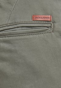 Primo piano di tessuto verde oliva con una tasca orizzontale e una piccola etichetta in pelle marrone cucita sopra, impressa con "JACK&JONES".