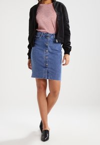 Kvinna klädd i en knappradad denimkjol, rosa skjorta, svart bomberjacka och svarta pumps, stående med benen korsade.