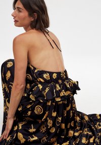 Robe maxi en tissu noir ornée de motifs dorés de différentes formes et designs. Dos nu avec une taille à volants et des bretelles à nouer.