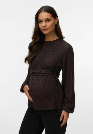 Zwangere vrouw met lang donker haar draagt een donkerbruine top met lange mouwen en zwarte broek, houdt voorzichtig haar buik vast.