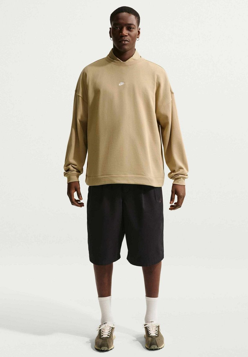 Uomo in piedi che indossa una felpa beige a maniche lunghe con il logo Nike, pantaloncini neri, calzini bianchi e sneakers marroni su uno sfondo bianco semplice.