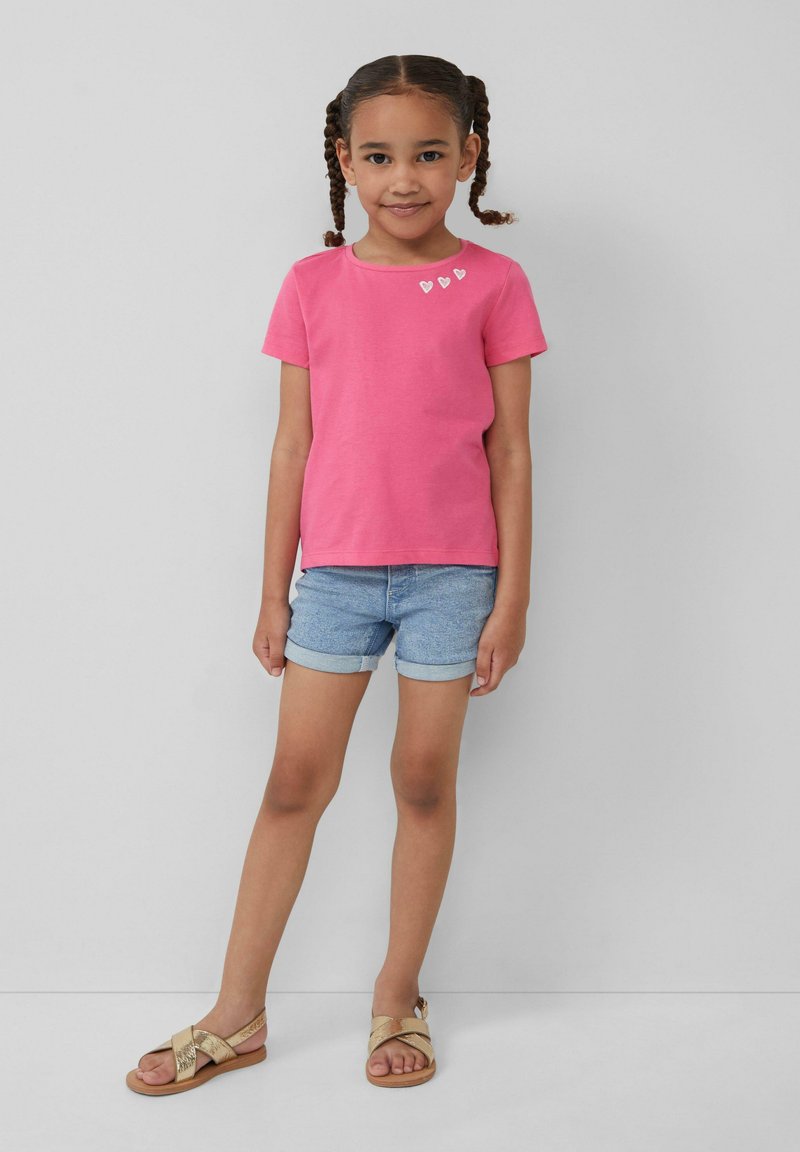 Roze t-shirt met korte mouwen met drie kleine witte hartontwerpen bij de halslijn, gecombineerd met lichte denimshorts en gouden sandalen.