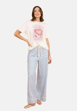 Femme portant un t-shirt graphique crème avec un design de gâteau et un pantalon large à carreaux bleus avec des motifs de cœurs rouges, debout pieds nus.